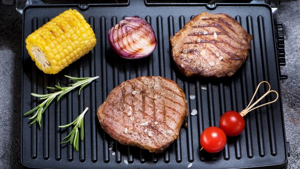 Image of electric grill with a number of different foods: maríz, cherry tomatoes, onion and meats for the grill.